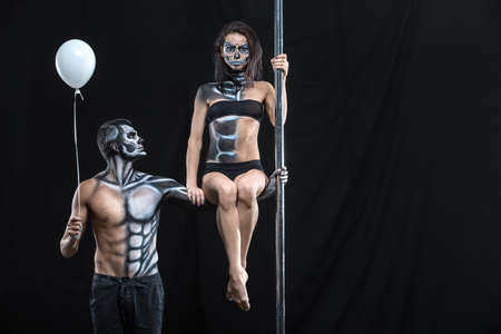 Sportive couple of pole dancers in the dark studio. Man holds a pylon with the left hand and holds a balloon in the right. Girl sits on the guy's left arm. They have a horrific body-art. Horizontal.の写真素材