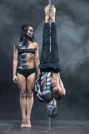Fabulous couple of pole dancers with a horrific body-art in the dark studio with a cloud of a smoke. Man hangs upside down on a pylon. Girl stands on her toes next to him. Vertical.の写真素材
