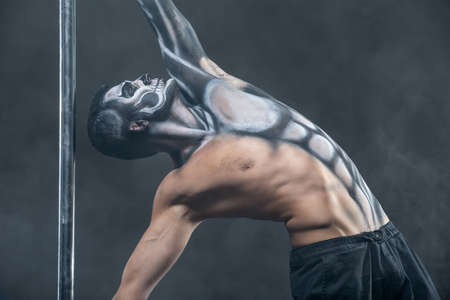 Strong pole dancer with horrific body-art next to a pylon in the dark studio with a smoke on the...