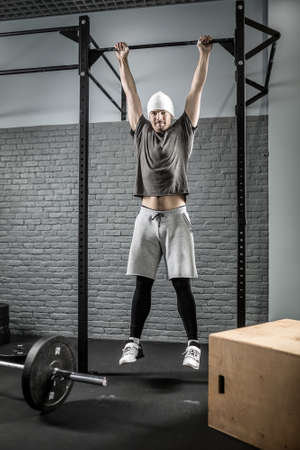 Young man with a beard hangs on a crossbar in the gym on the gray brick wall background. He wears...