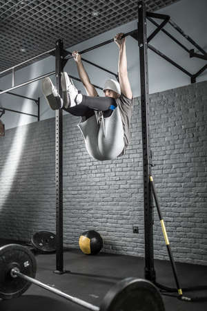 Strong man pulls up his legs while hangs on a crossbar in the gym on the gray brick wall background....