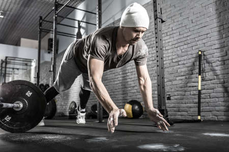 Cool guy with a beard makes a pushup with a front clap in the gym on the gray brick wall background. He wears sportswear, white sneakers and a white cap. Man looks in front of himself. Horizontal.の写真素材
