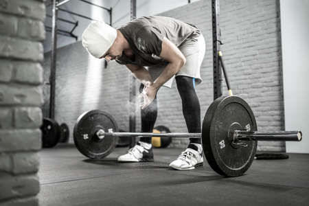 Healthy guy with a beard tilts to a barbell in the gym on the gray brick wall background. He wears sportswear, white sneakers and a white cap. Beam of light falls on the powder cloud near his hands.の写真素材