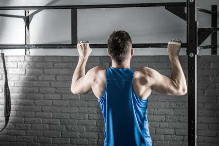 Active man makes a pull-up on the crossbar in the gym on the gray brick wall background. He wears a...