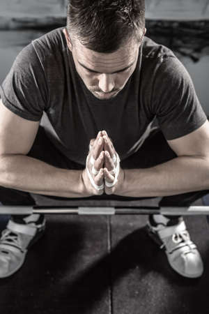 Peaceful man with a beard sits on the floor above the chrome barbell bar in the gym. He holds his hands together. There are plasters on his fingers and hands. Guy wears sportswear with white sneakers.の写真素材