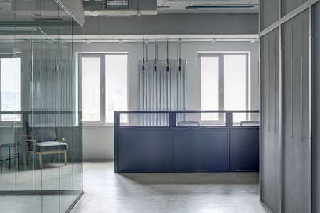 Business interior in a loft style with gray walls. There is a blue metal reception rack with two armchairs on the windows background, work zones with glass and mesh partitions. Horizontal.の写真素材