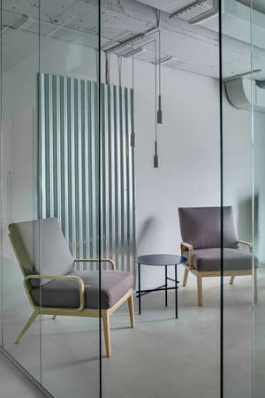 Armchairs with wooden legs and a small round metal table on the background of the gray wall with the metal panel. In front of them  there is a glass partition with an entrance. Indoors. Vertical.の写真素材