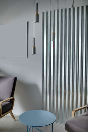 Small blue round metal table between two armchairs on the background of the metal panel on the gray wall. Three lamps are hanging on the ceiling. Indoors. Vertical.の写真素材