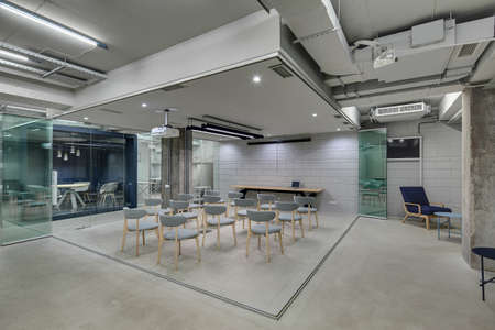 Presentation zone in the office in a loft style with brick walls and concrete columns. Zone has many gray chairs and a projector above them. On the left there is blue meeting zone with glass doors.の写真素材