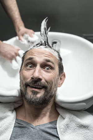 Funny man with a beard lies on the white sink in the barbershop. He has the lathered head with the punk hairstyle. Guy looks to the side with a grimace. Closeup. Vertical.の写真素材