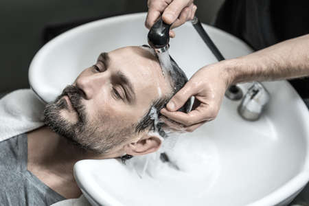 Cute man with a beard lies on the white sink in the barbershop. Barber washes his head with the black faucet. Guy wears a gray T-shirt and a white towel. Closeup. Horizontal.の写真素材