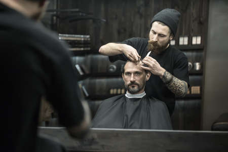 Stylish bearded man in a black cutting hair cape in the barbershop. Brutal barber with a tattoo in a black cap is combing his hair. Horizontal.の写真素材