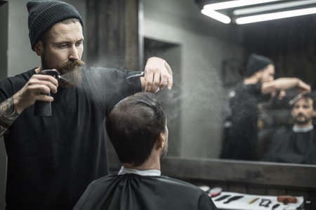 Attentive barber with a big beard splashes from the spray bottle at the hair of his client in the barbershop. He wears a black T-shirt with a black cap. They both reflected in a blurry mirror.の写真素材