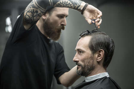 Tattooed barber with a big beard is cutting the hair of his client in the black cape in the barbershop. He wears a black T-shirt. Customer wears a black cutting hair cape with a neck paper.の写真素材