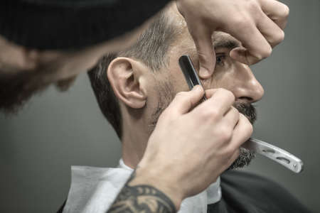 Elegance man with a beard in the barbershop. Barber with a tattoo is trimming his beard with a straight razor. Closeup. Horizontal.の写真素材