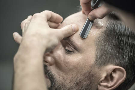 Bearded man in the barbershop. Barber is trimming his hair with a straight razor. Closeup....