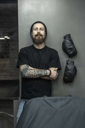 Brutal barber with a tattoo, a big beard and a mustache stands on the gray wall background in the barbershop. He wears light jeans, a black T-shirt and a cap and looks into the camera. Vertical.の写真素材