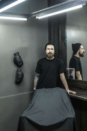 Trendy barber with a tattoo, a big beard and a mustache stands in the barbershop. He wears a black T-shirt and a cap and looks into the camera. Guy reflected in a mirror. Vertical.の写真素材