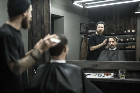 Intent barber with a big beard and a tattoo prepares to do a haircut for the stylish man in the barbershop. He wears a black T-shirt with a cap and holds a hair comb. They both reflected in a mirror.の写真素材