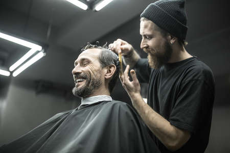 Laughing bearded man with closed eyes in the black cutting hair cape in the barbershop. He has hairgrips on his head. Pleased barber in a black T-shirt and a cap is doing the haircut. Horizontal.の写真素材