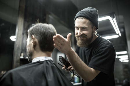 Happy barber with a beard looks at the hair of his client in the barbershop. He holds a cutting comb and a hair clipper. Hairdresser wears a black T-shirt and a cap. Horizontal.の写真素材