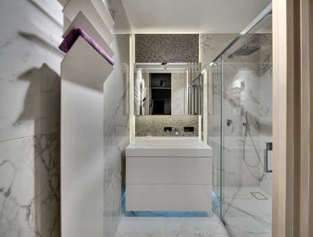 Bathroom in a modern style with light tiles on the walls and the floor. There is a white sink  with a chrome faucet and a mirror, a shower with glass door, a white stand with a towel.の写真素材