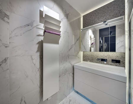 Modern bathroom with light tiles on the walls and the floor. There is a white sink  with a chrome faucet and a mirror, a white stand with a towel. Over the sink there is a mosaic wall. Horizontal.の写真素材