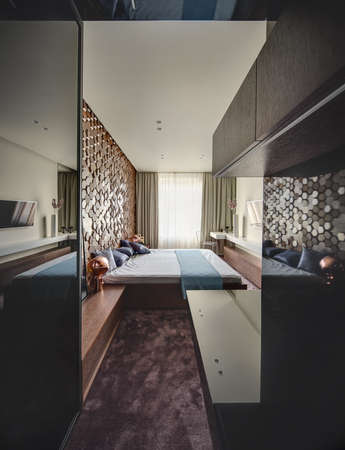 Bedroom in a modern style with burgundy carpet on the floor. There is a bed with pillows and a coverlet, TV, design lamps, white rack with a vase with a flower, chair. Wall next to bed is textured.の写真素材