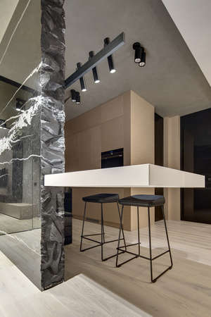 Light tabletop and a black marble wall with white patterns in the kitchen zone in a modern style. There are two black stools under the tabletop. Behind them there are wooden lockers with an oven.の写真素材