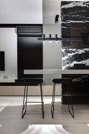 Black marble wall with white patterns with a light tabletop and two black stools under it. There are glowing lamps, white wall with a TV, decorations, black fireplace with conditioner, wooden lockers.の写真素材