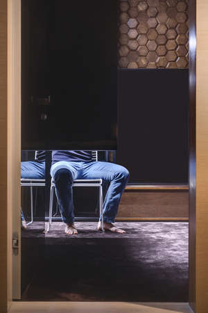 Hall in a modern style with a burgundy carpet on the floor and a wooden wall textured with hexagons. There is an open door of the specular wardrobe, barefoot man sitting on a white chair, wooden rack.の写真素材