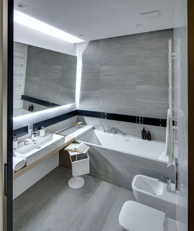 Bathroom in a modern style tiled with the white and gray tiles. There is a white sink with accessories, mirror, bath, towel holder, towel radiator, toilet and a bide, stand with a towel and a book.の写真素材