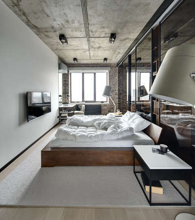 Bedroom in a loft style with brick wall and concrete ceiling. There is a TV, bed with pillows, lamps with lampshades, wardrobe with glass sliding doors, tables, armchair, carpet on the floor.の写真素材