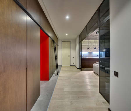 Hall in a modern style with light walls, glass partitions and partially concrete ceiling. There are wooden lockers and wardrobes, ladder, door, sofa, glowing kitchen zone with hanging lamps.の写真素材