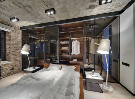 Loft style bedroom with brick wall and concrete ceiling. There is a bed with white pillows and blankets, glowing lamps, wardrobe with glass sliding doors, tables, parquet and a carpet on the floor.の写真素材