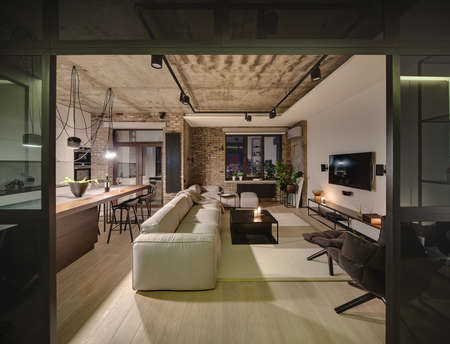 Luminous sitting-room in a loft style with different walls and a concrete ceiling. There are light sofas, wooden tables, different chairs, armchair, TV, lamps, kitchen zone with kitchen equipment.の写真素材