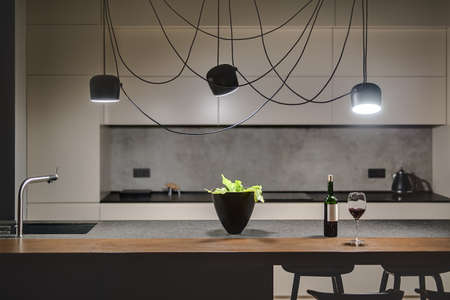 Loft style kitchen with a concrete wall. There is a kitchen island with a sink, plant, bottle and glass, black chairs, light lockers, dark tabletop with stove and teapot, hanging black glowing lamps.の写真素材