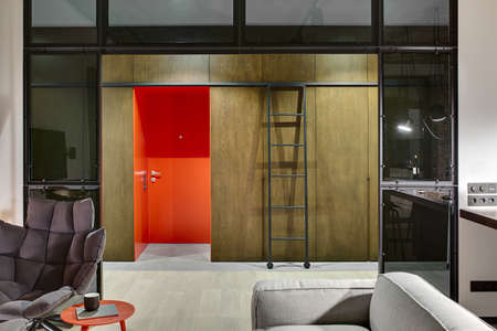 Luminous hall in a loft style with a red entrance door, wooden wardrobes and lockers, ladder with wheels, armchair, table with book and cup, sofa, glass partitions. Parquet and tiles are on the floor.の写真素材