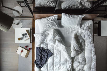 Top view at the bed with white pillows and blankets and blue T-shirt. There are white tables, lamp...