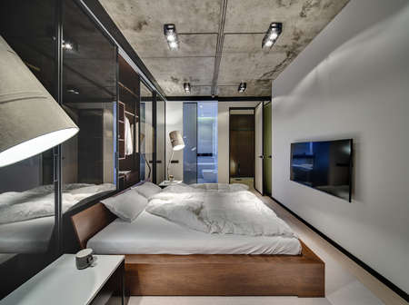 Room in a loft style with white walls and concrete ceiling. There is a TV, bed with pillows and blankets, glowing lamps, wardrobe with glass sliding doors, tables, doors to the bathroom and corridor.の写真素材