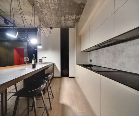 Kitchen zone in a loft style with a parquet on the floor. There is a kitchen island with sink and faucet, bottle and glass, black chairs, light lockers, dark tabletop with a stove, glowing lamps.の写真素材