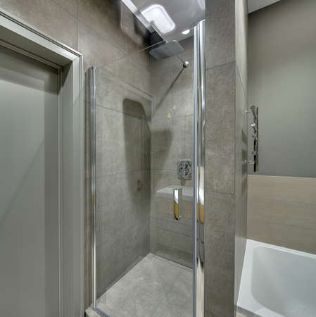 Contemporary bathroom with long tiles on the walls and floor. There is a shower cabin with a glass door, white bath, door, chrome towel holder, glowing lamps. Vertical.の写真素材