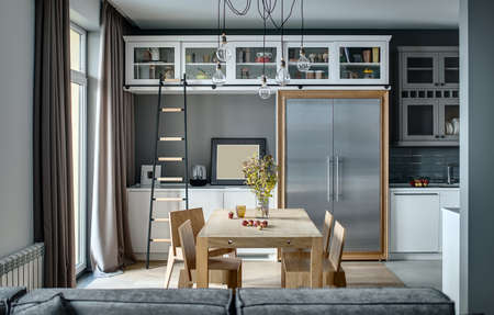 Kitchen in a modern style with gray walls, white lockers and shelves with accessories. There is a wooden table with chairs, dark ladder, fridge with metal doors, sofa, sink with faucet, hanging lamps.の写真素材