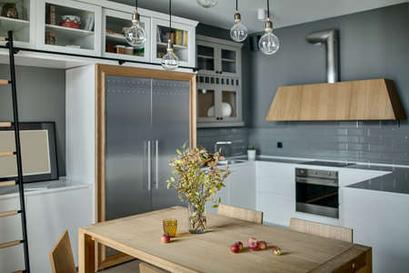 Cool kitchen in a modern style with gray walls, white lockers and shelves with accessories. There is a wooden table with branches in a vase and chairs, ladder, fridge, sink, oven, stove, kitchen hood.の写真素材