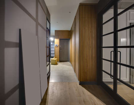 Hallway in a modern style with glowing lamps and gray walls. On the floor there are tiles and a parquet. There is a large wooden wardrobe, gray door, yellow pouf and glass black doors. Vertical.の写真素材