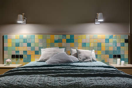 Wide bed with many pillows and a coverlet on the background of the colorful tiles with black switches and power sockets. There are wooden nightstands with flower and candles, glowing white lamps.の写真素材
