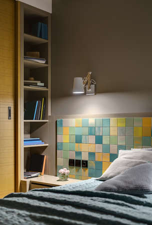 Bed with several pillows and a coverlet on the background of the colorful tiles with black switches and a power socket. There is a wooden nightstand with flower, shelves with books, glowing lamp.の写真素材