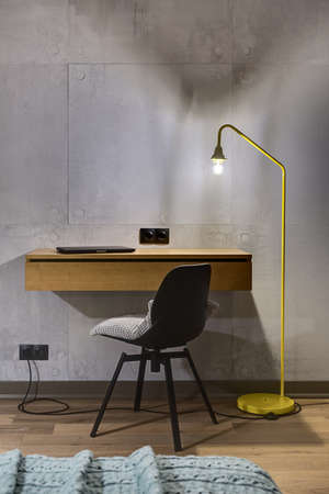 Wooden rack with a laptop and a black chair with a gray pillow on the background of the gray tiled wall. Next to the rack there is a glowing trendy yellow lamp. Black power sockets are on the wall.の写真素材