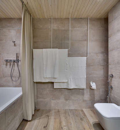 Modern bathroom with textured tiles and a wooden ceiling. There is a white bath with a curtain and a shower, towel holders, toilet with another shower, toilet paper holder. Horizontal.の写真素材