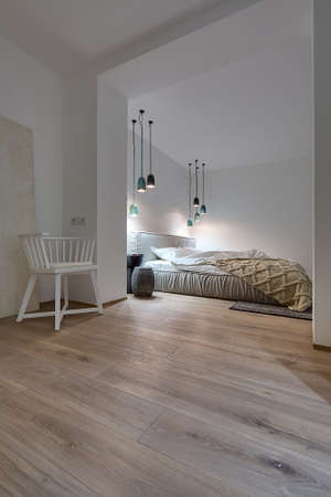 Modern bedroom with white walls and a parquet with a carpet on the floor. There is a bed with a white pillows and blanket and brown plaid, nightstand with a book and a lampshade, glowing lamps, chair.の写真素材